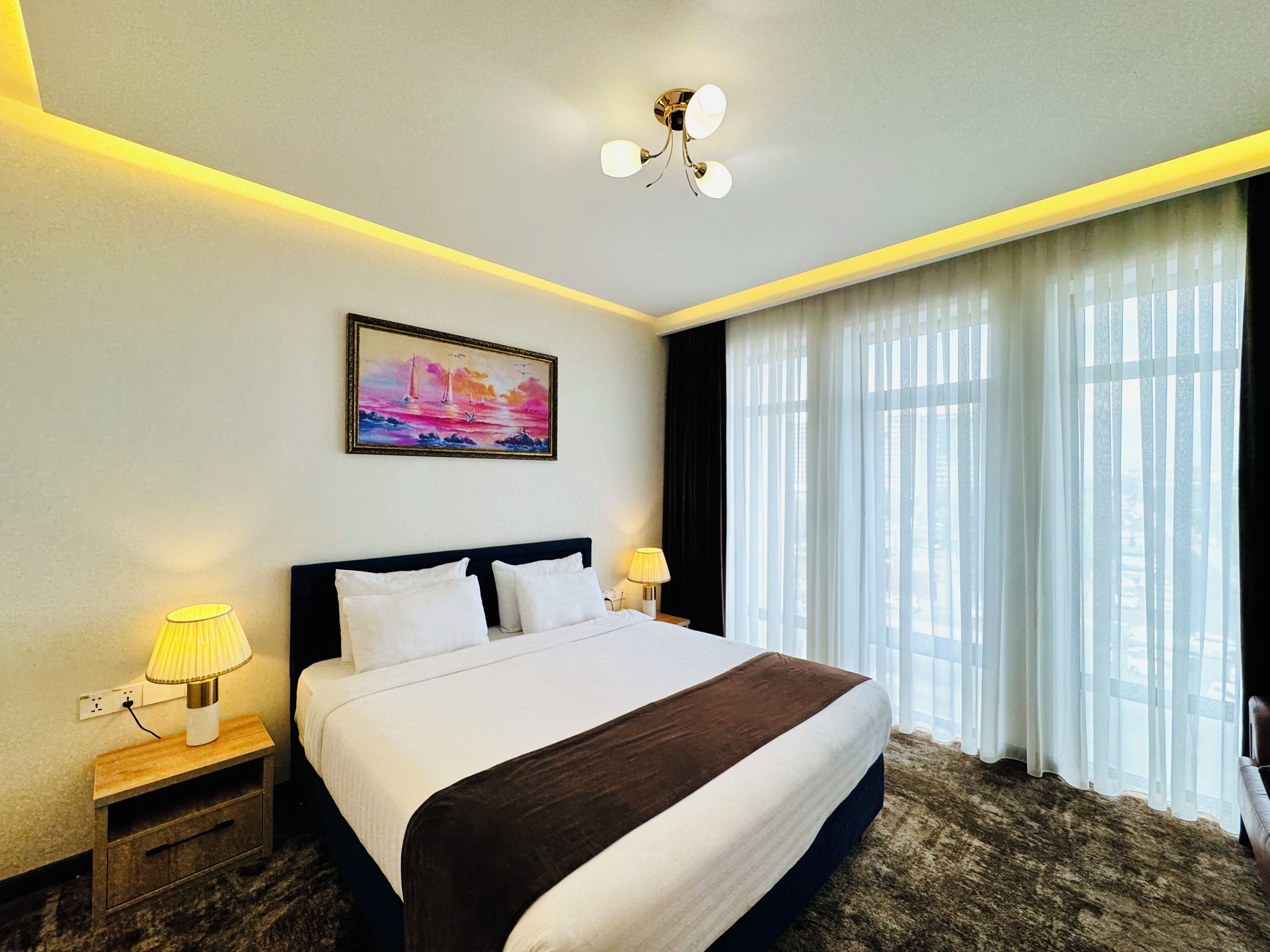 Habitación de hotel con una cama grande, una mesita de noche con lámpara, un cuadro en la pared y vistas a través de cortinas traslúcidas.