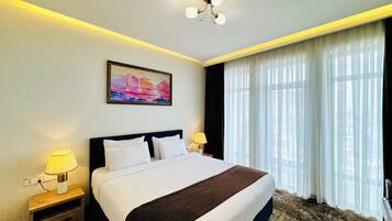 Habitación de hotel con una cama grande, una mesita de noche con lámpara, un cuadro en la pared y vistas a través de cortinas traslúcidas.