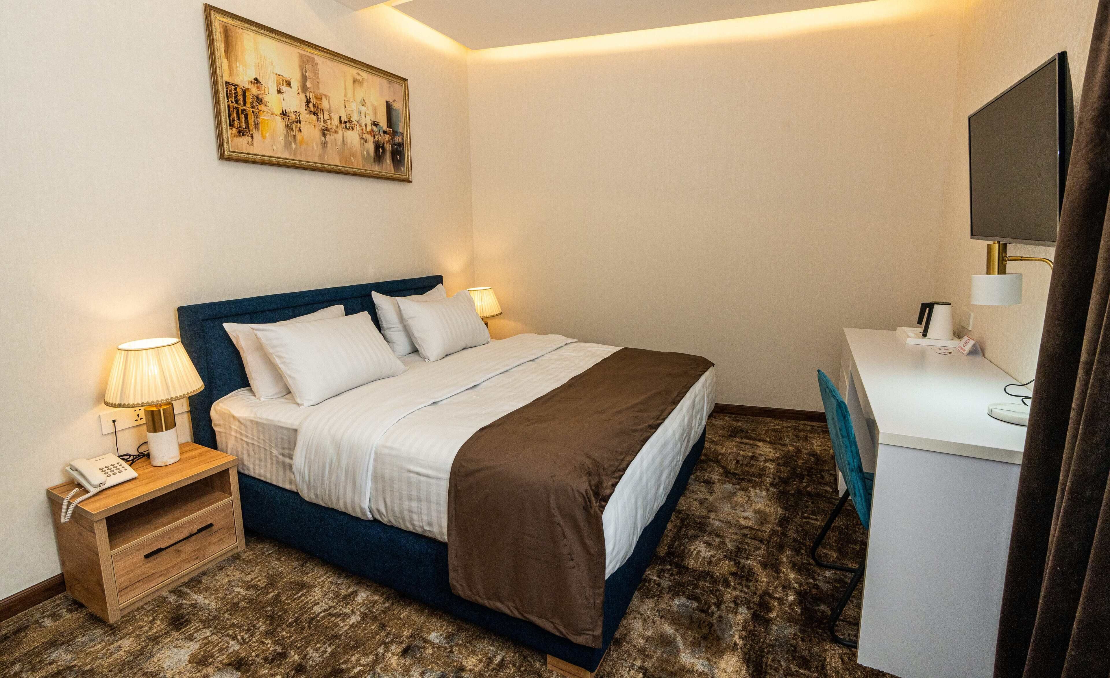 A hotel room with a bed, bedside lamps, a desk with a computer, and a painting on the wall.