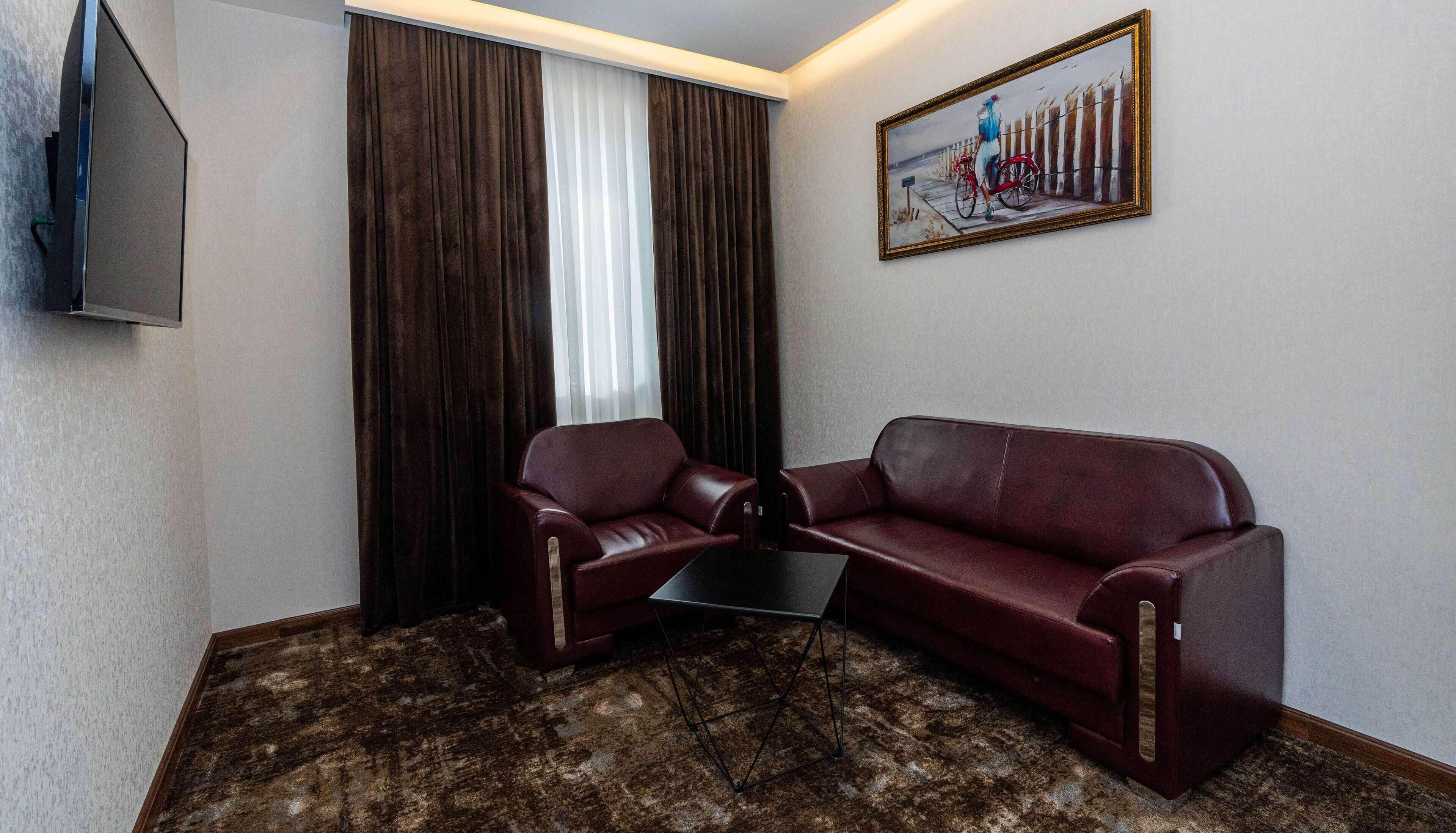Una habitación de hotel con alfombra de color marrón oscuro y diseño, un sofá de cuero rojo, un sillón rojo de cuero más pequeño, una mesa de centro negra y un cuadro de una mujer en bicicleta.