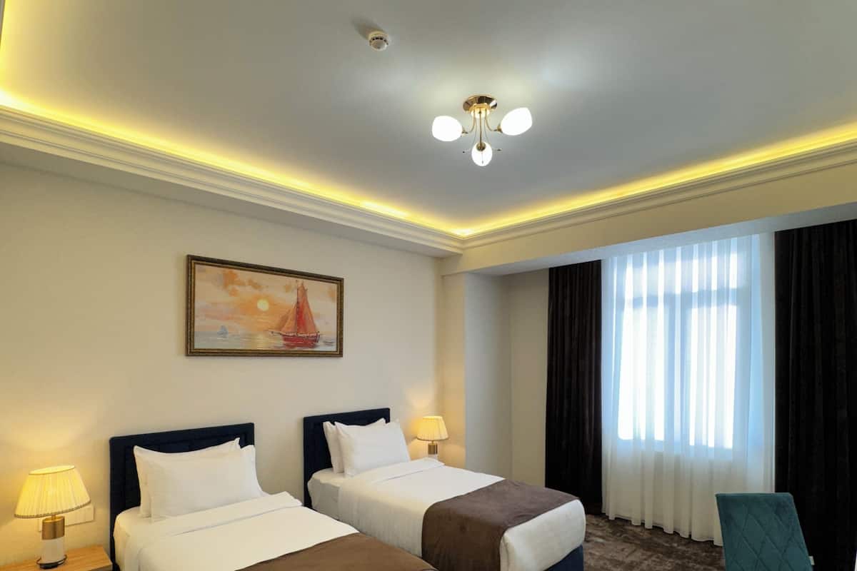 Une chambre d’hôtel avec deux lits, un tableau au mur, une fenêtre avec des rideaux, une chaise et un luminaire au plafond.