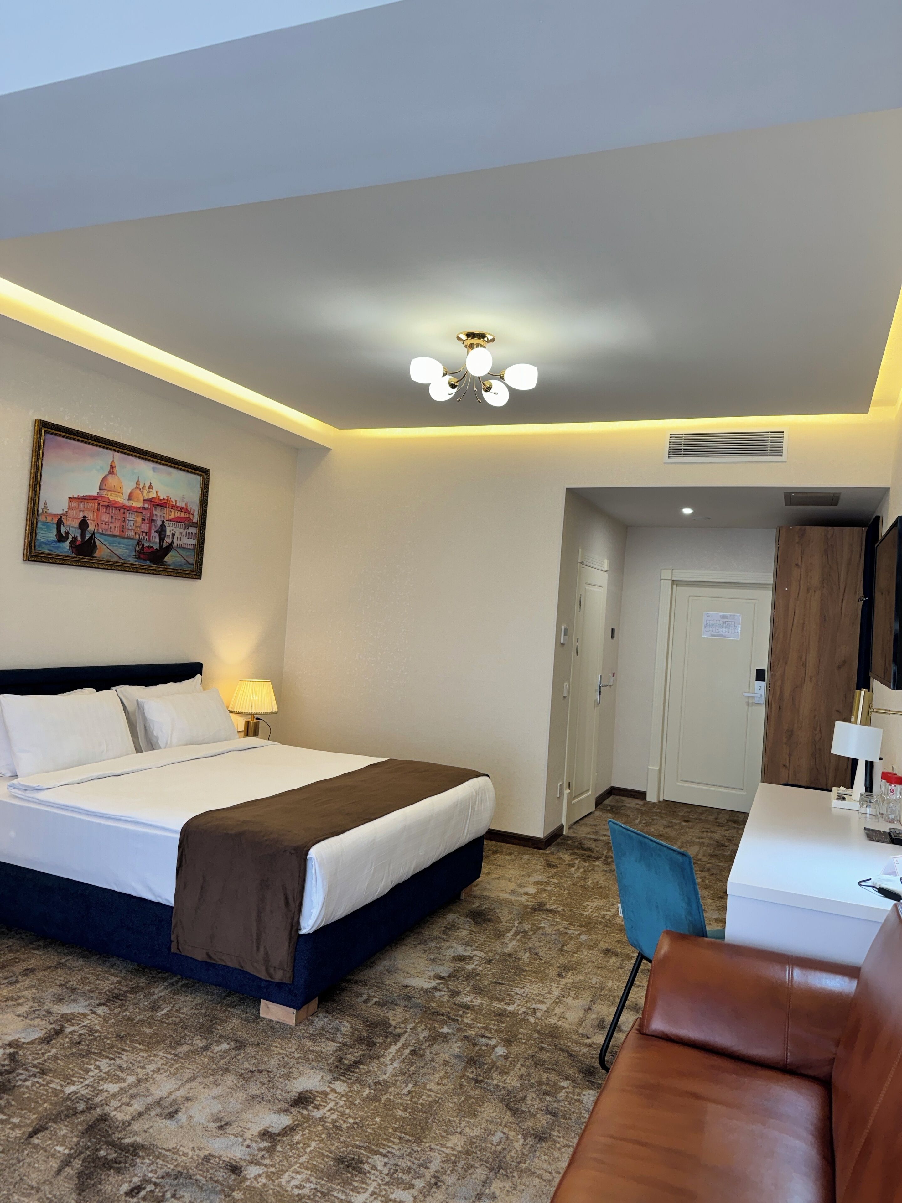 A hotel room with a bed, a desk, a chair, a painting of Venice, and a ceiling light.