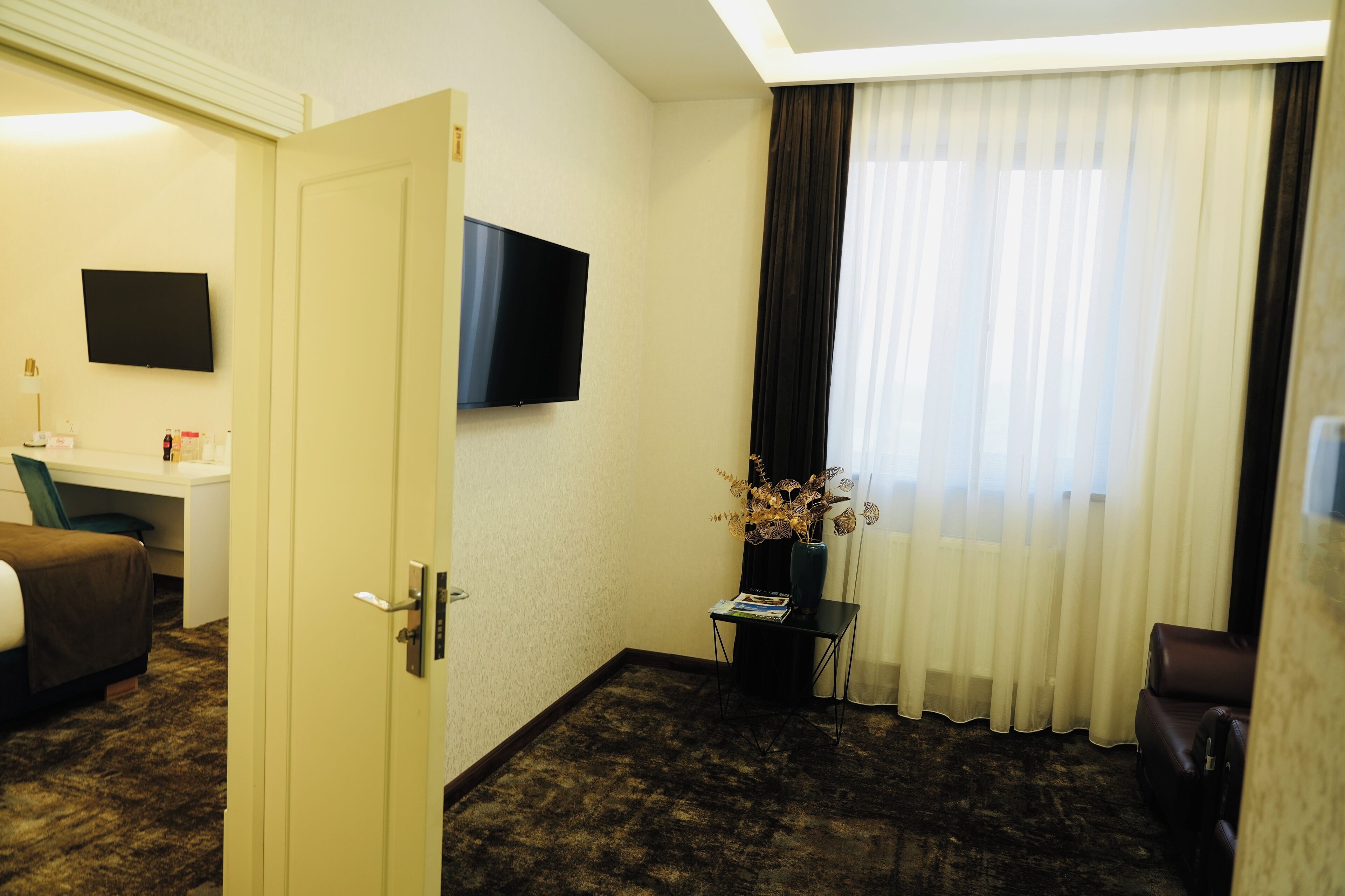 A hotel room with a bed, a desk, a TV, a window with curtains, and a door.