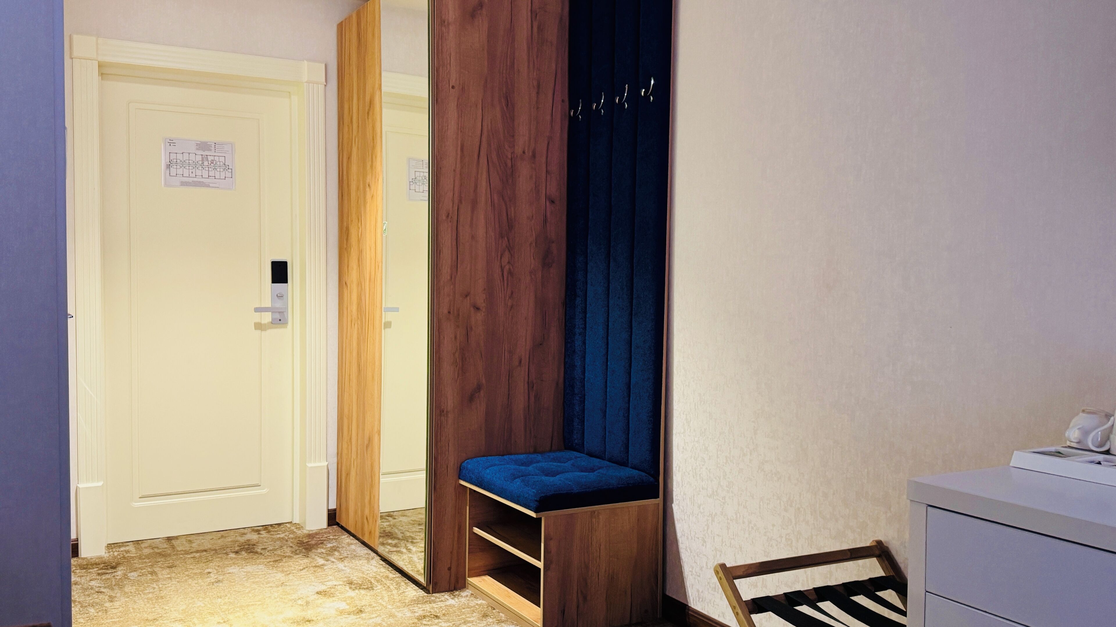 A narrow hallway with a white door, a wooden bench with a blue cushion, and a small desk with a white surface.