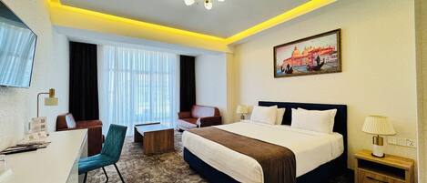 A hotel room with a bed, two chairs, a desk, a TV, and a painting of a cityscape.