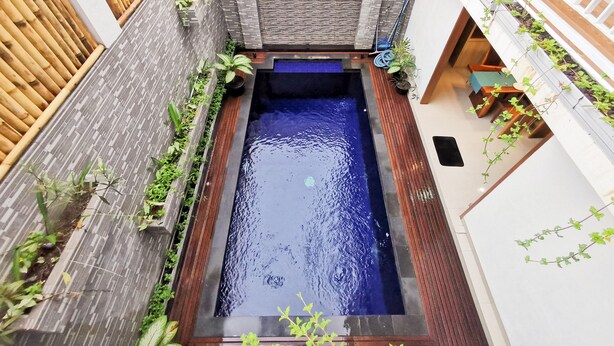 A rectangular pool with a dark blue water feature, surrounded by a wooden deck and a brick wall with greenery. - Sunny Apartico Villa (Ungasan)