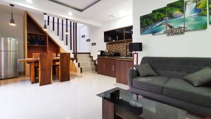 A modern living room with a sofa, a glass coffee table, a dining area with a wooden table and chairs, a kitchen with a black countertop, and a staircase with wooden railings. - Sunny Apartico Villa (Ungasan)