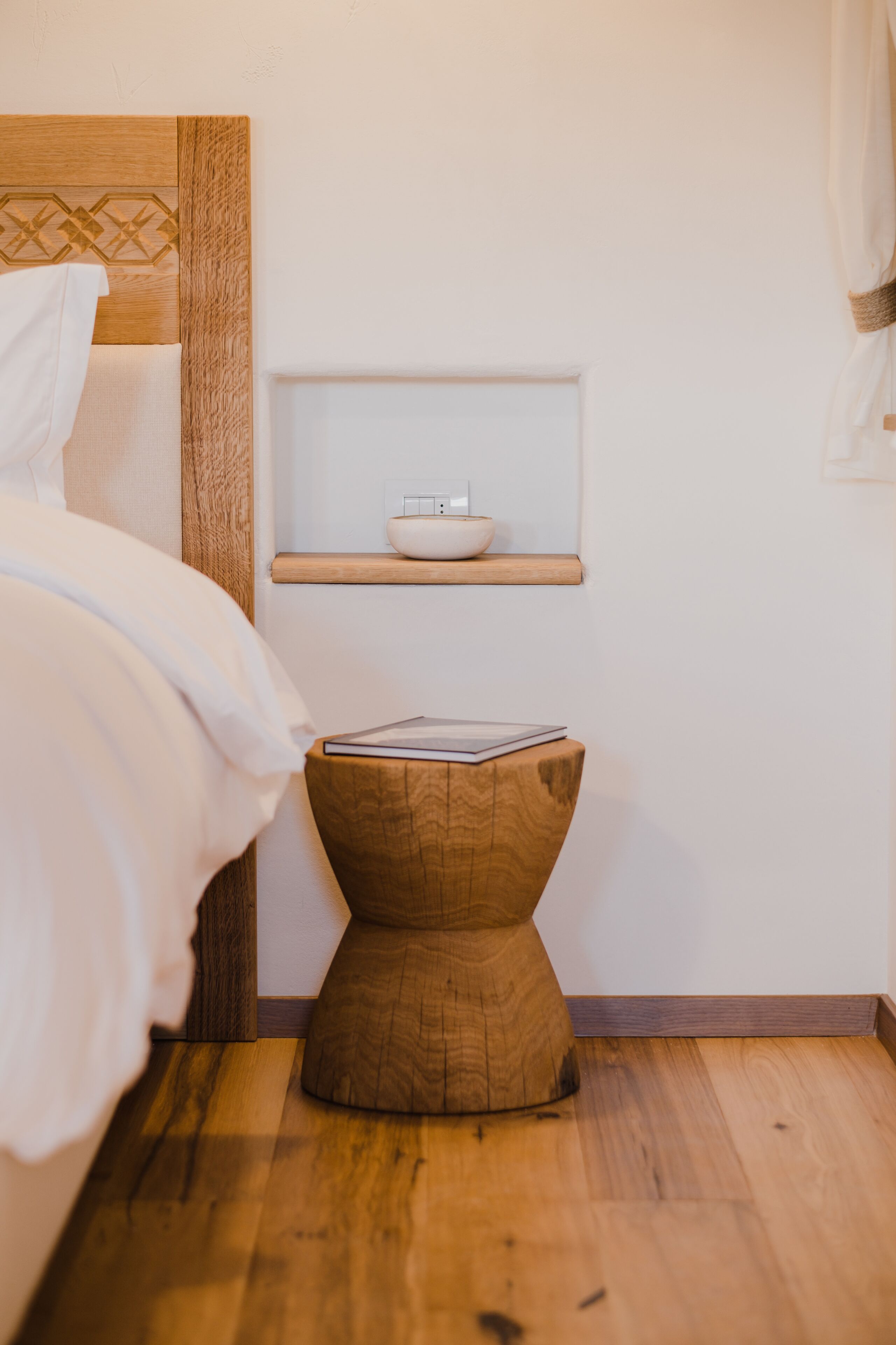 Un dormitorio con cabecera de madera, una mesita de noche con un libro y un pequeño estante con un cuenco.