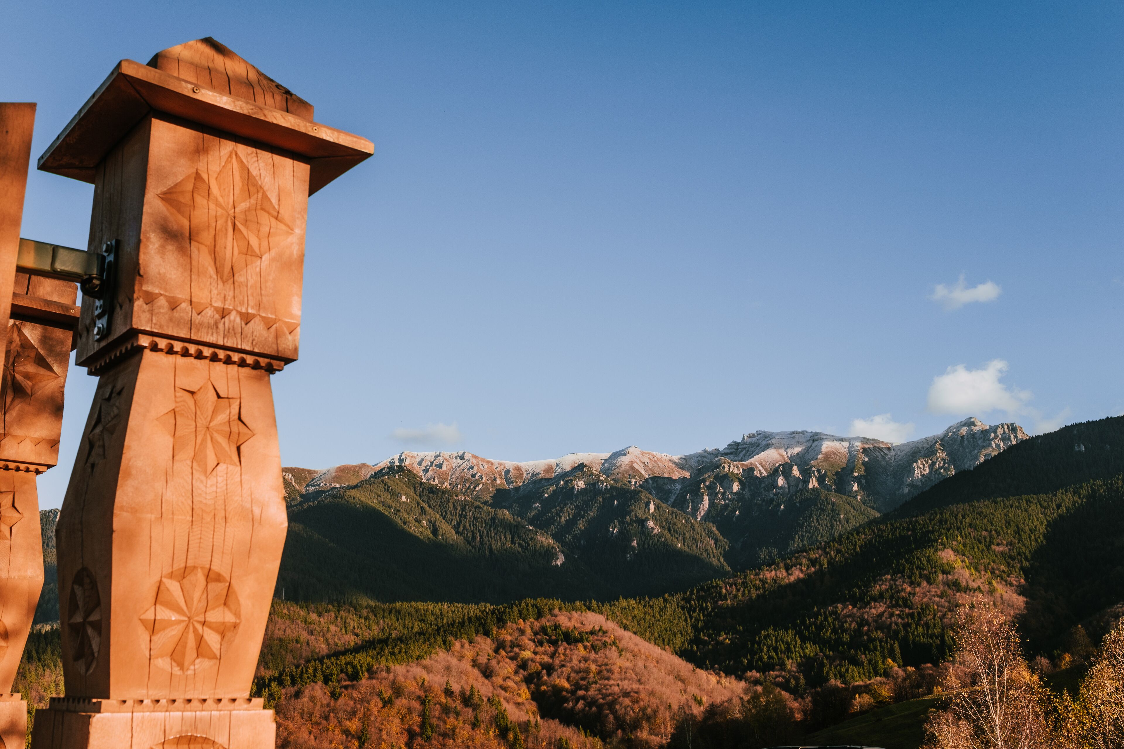 A wooden structure with carved geometric patterns, set against a mountainous landscape with autumn foliage and clear skies.