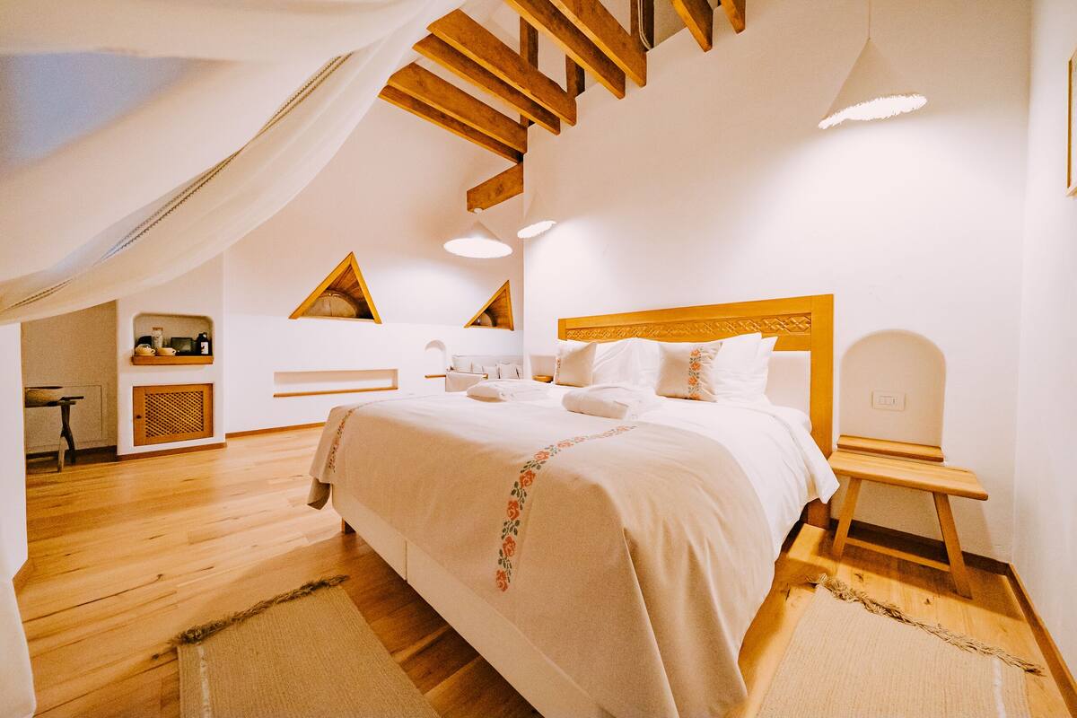 Une chambre avec un grand lit, des poutres apparentes, une lucarne et un petit bureau.