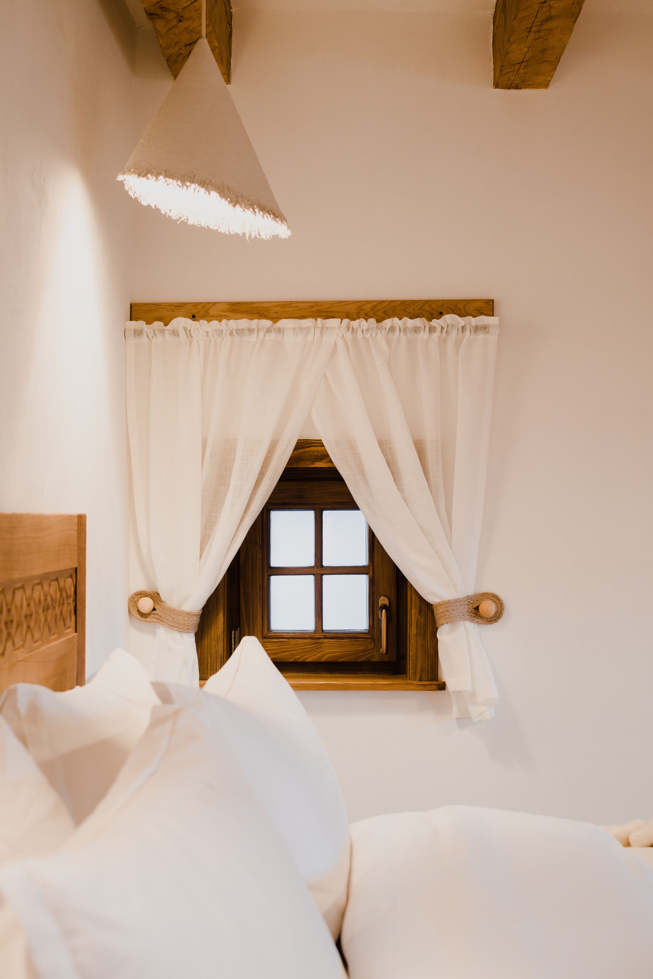 A bedroom with a wooden headboard, a window with white curtains, and a hanging light fixture.