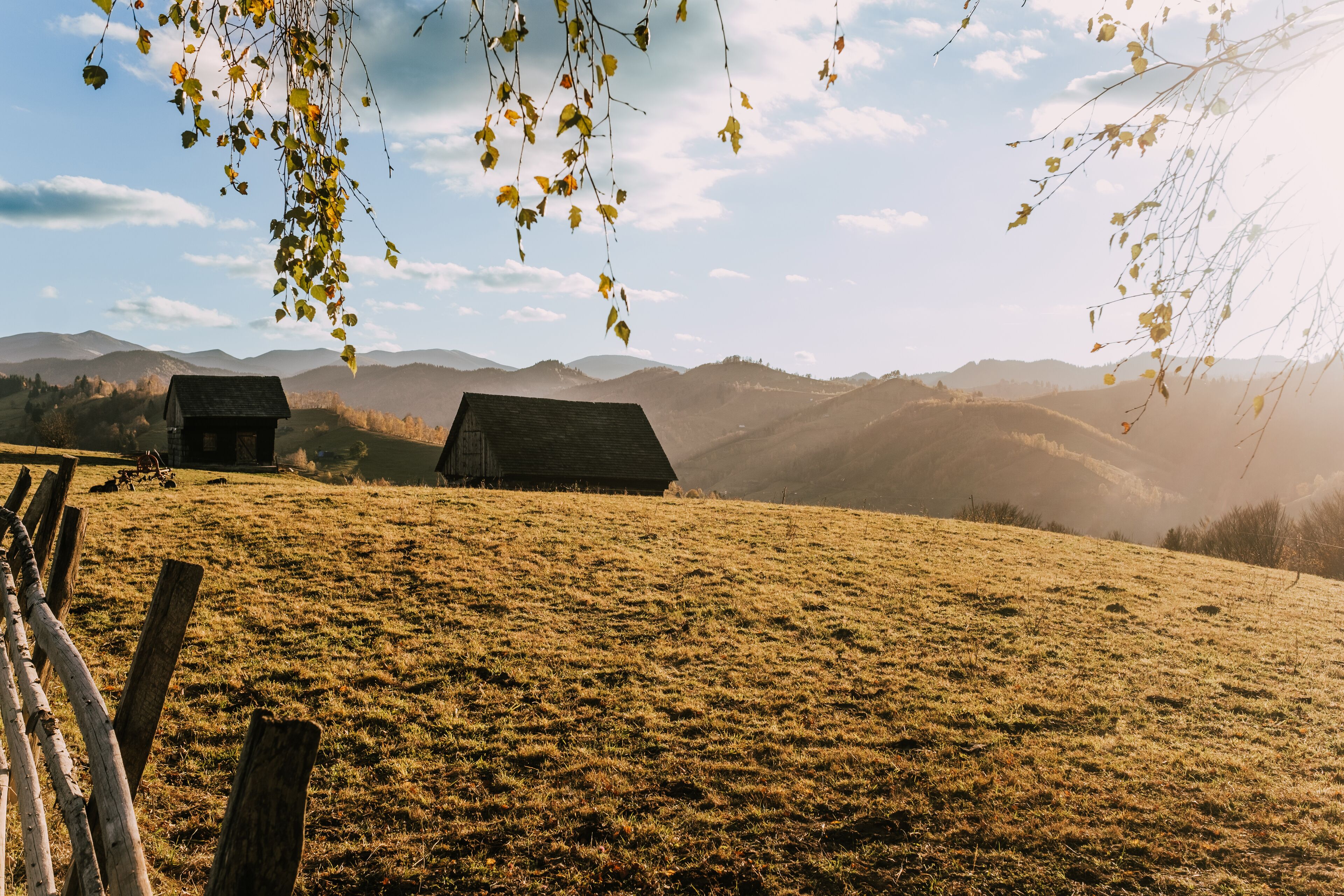Un paisaje rural con dos cabañas de madera en una colina cubierta de hierba, rodeado de montañas.