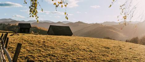 Un paisaje rural con dos cabañas de madera en una colina cubierta de hierba, rodeado de montañas.