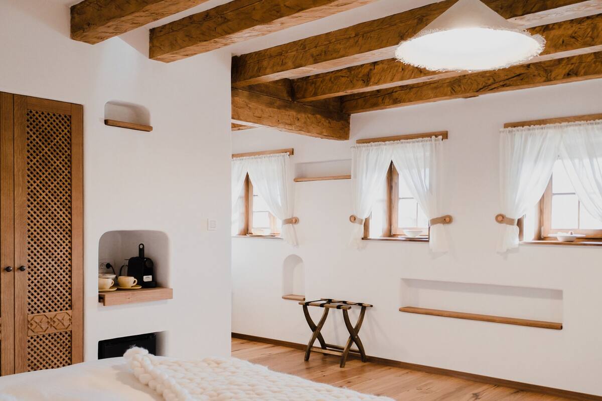 Une chambre avec un grand lit, des poutres en bois, un luminaire suspendu et une fenêtre avec des rideaux.