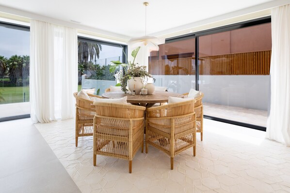 A dining area with a round table and four wicker chairs, a potted plant, and a view of the outdoors through large windows.