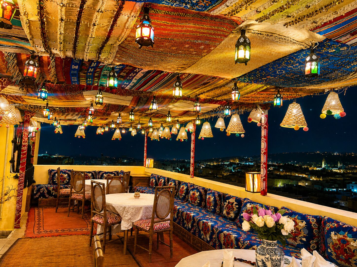 Un espacio de comedor al aire libre con vistas a la ciudad de noche, con faroles tradicionales y textiles estampados.