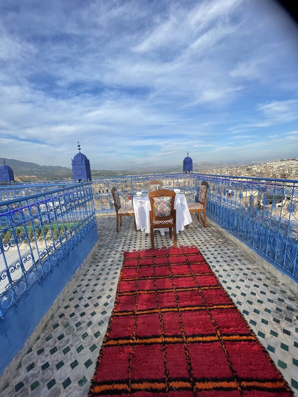 A terrace with a table set for two, a red patterned rug, and a view of a cityscape with distant mountains. - Riad Fes Palais Bab Sahra & Cooking Workshops  (Fes)
