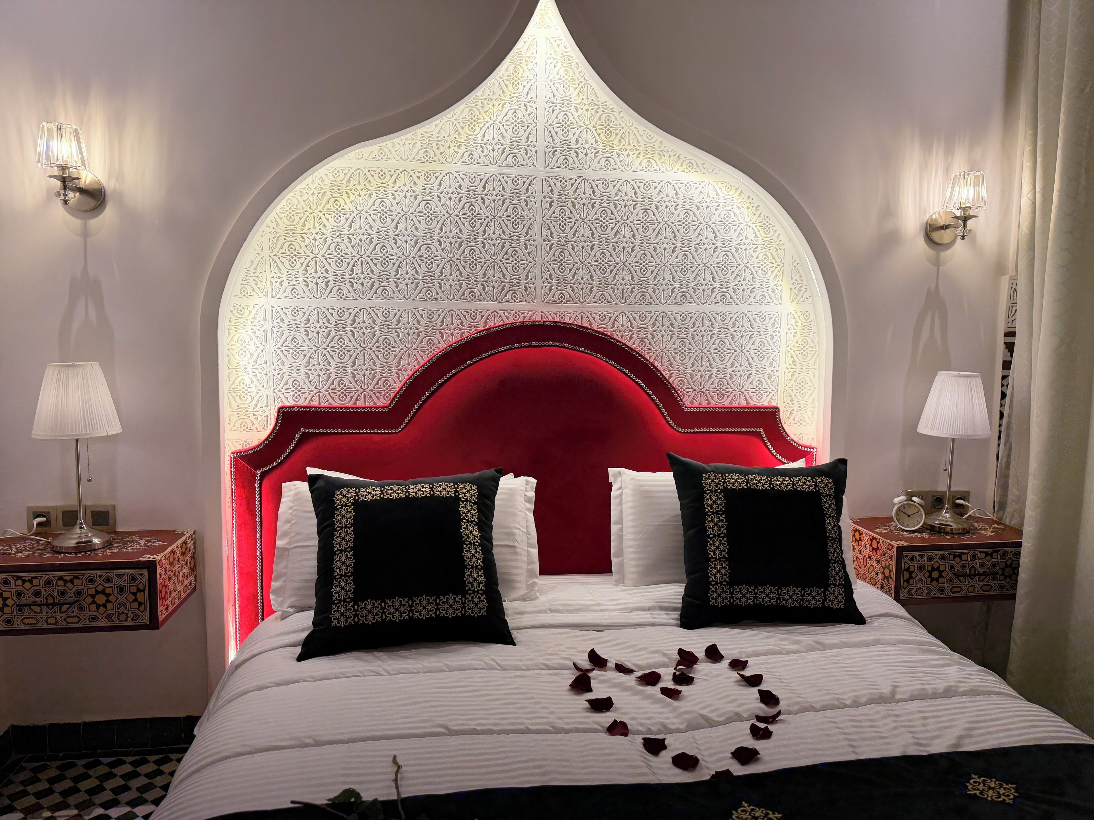 A hotel room with a red upholstered headboard, white patterned wall, and two bedside tables with lamps.