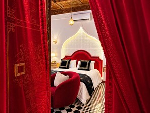 Une pièce avec un tapis à motifs, un fauteuil recouvert de tissu rouge, un lit avec une tête de lit blanche et un luminaire suspendu.