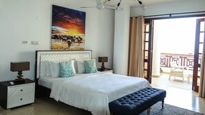 A bedroom with a large bed, two bedside tables with lamps, a bench at the foot of the bed, a ceiling fan, and a balcony with a railing.
