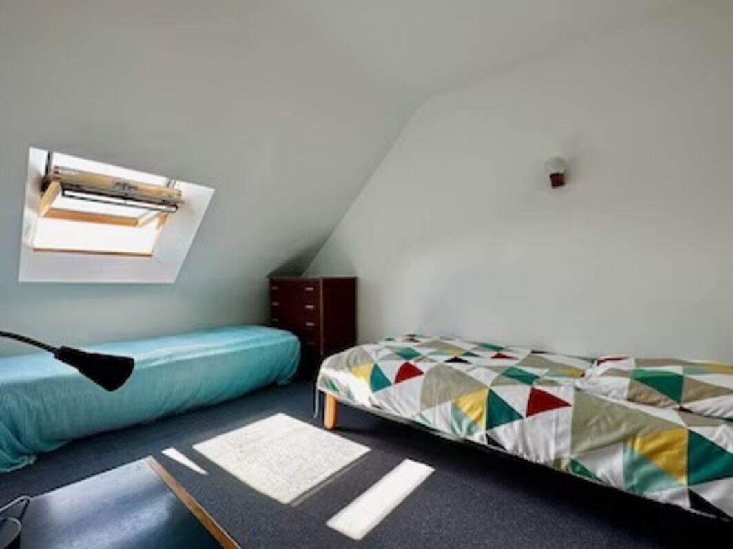Ein kleiner, gemütlicher Raum mit schrägen Wänden, einem Bett mit bunter Steppdecke, einem Schreibtisch mit Lampe und einem Fenster, das natürliches Licht hereinlässt.