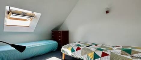 Ein kleiner, gemütlicher Raum mit schrägen Wänden, einem Bett mit bunter Steppdecke, einem Schreibtisch mit Lampe und einem Fenster, das natürliches Licht hereinlässt.