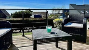 Un espace extérieur aménagé en patio, avec une table et deux chaises en osier, un verre contenant une boisson, et une clôture en métal en arrière-plan.