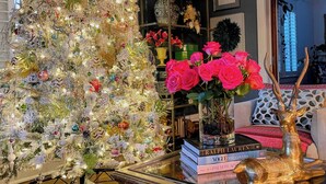 Uma árvore de Natal decorada com luzes e enfeites, um vaso de rosas cor-de-rosa e uma estante com livros e objetos decorativos.