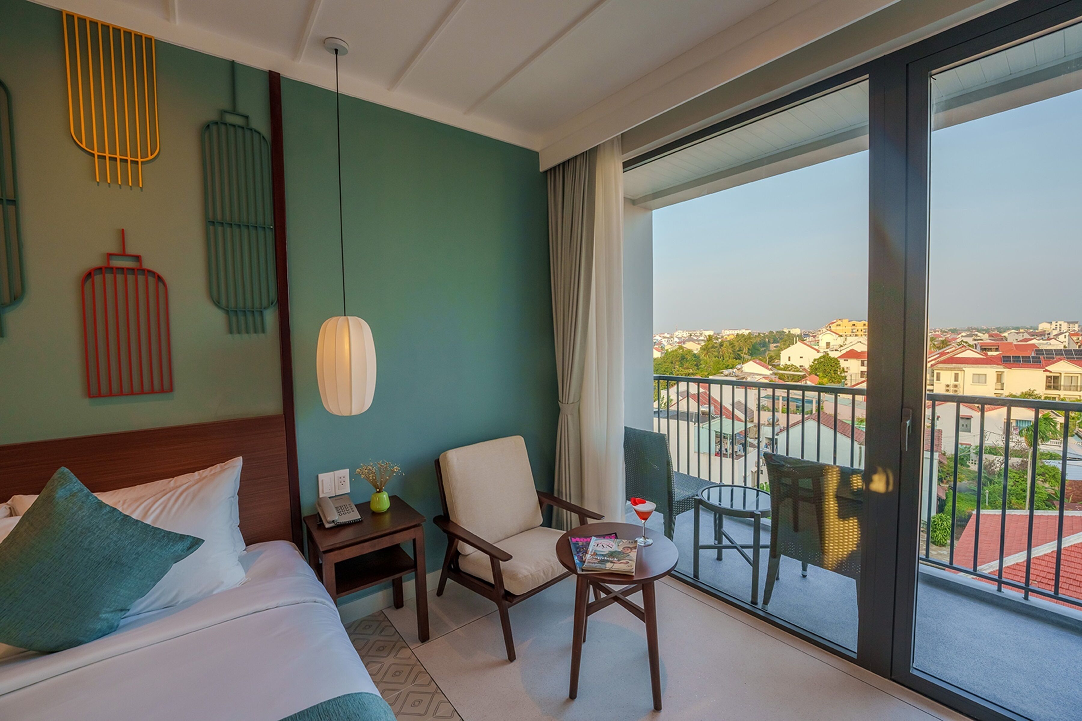 A hotel room with a bed, a chair, a small table, a nightstand, and a balcony with a view of the city.