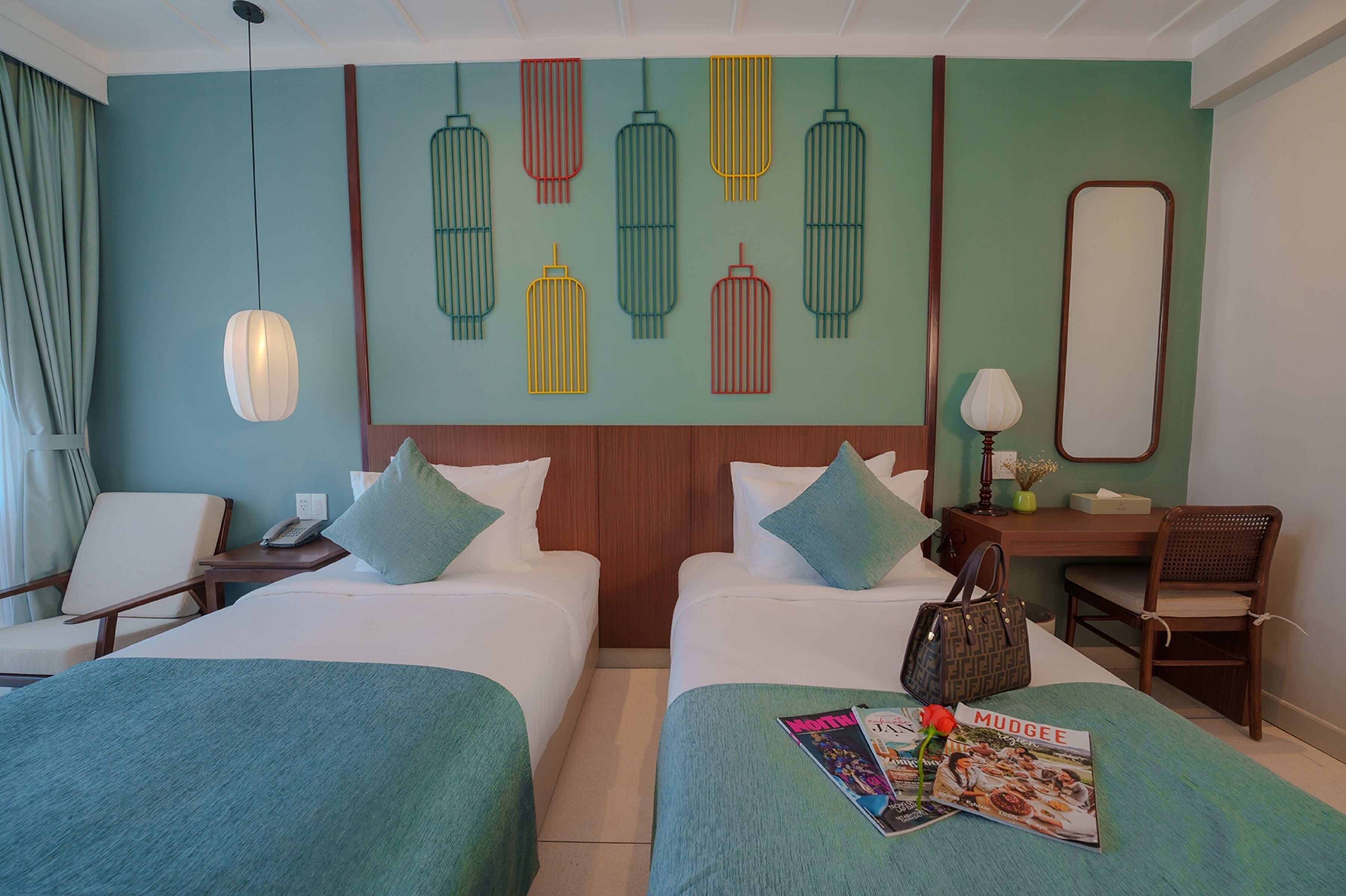 A hotel room with two beds, a desk, a chair, a lamp, and a wall with decorative hanging elements.