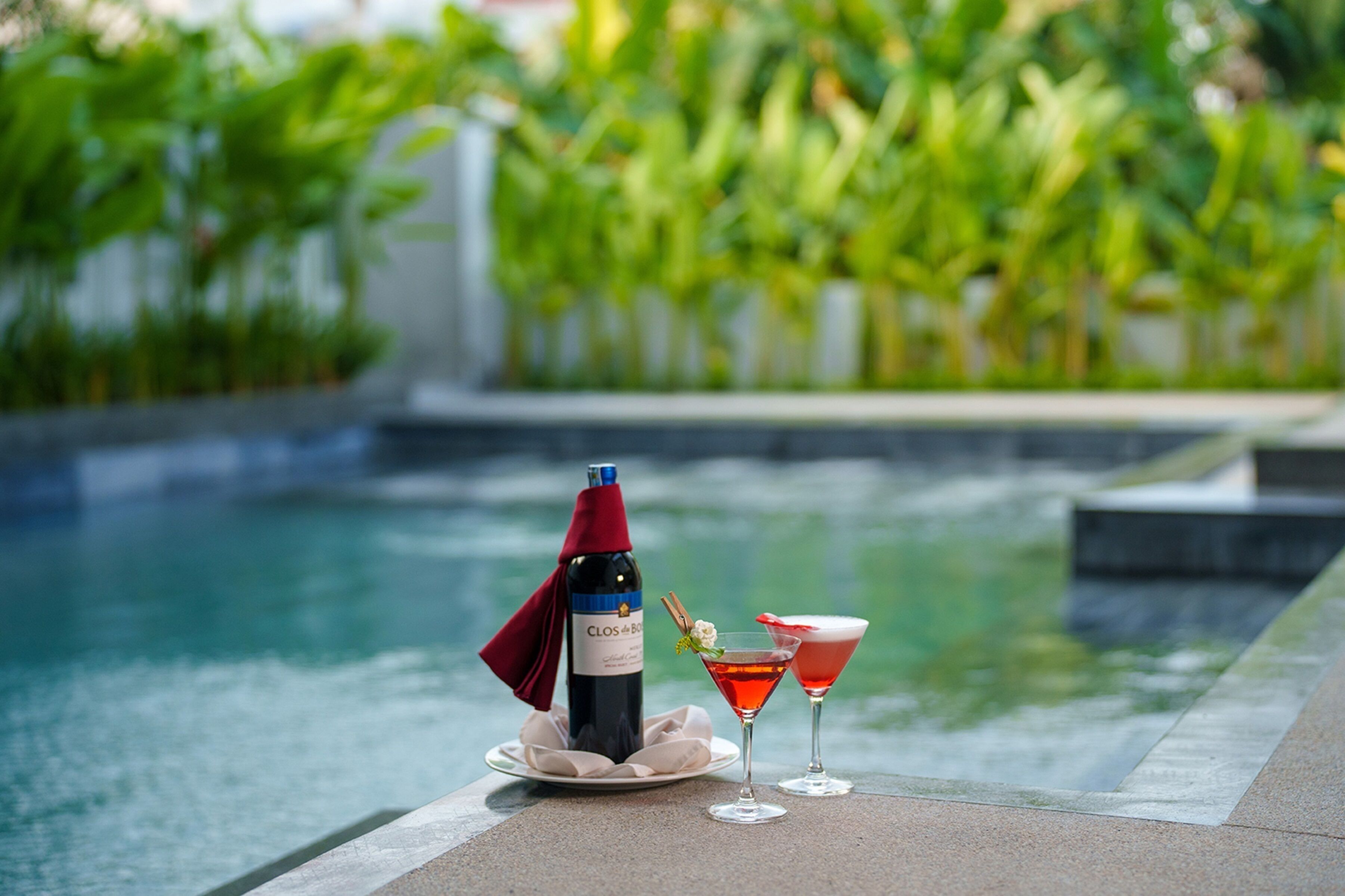 Una botella de vino Clos & Bock y dos copas de vino tinto sobre una mesa junto a la piscina, con vistas a una piscina y follaje.