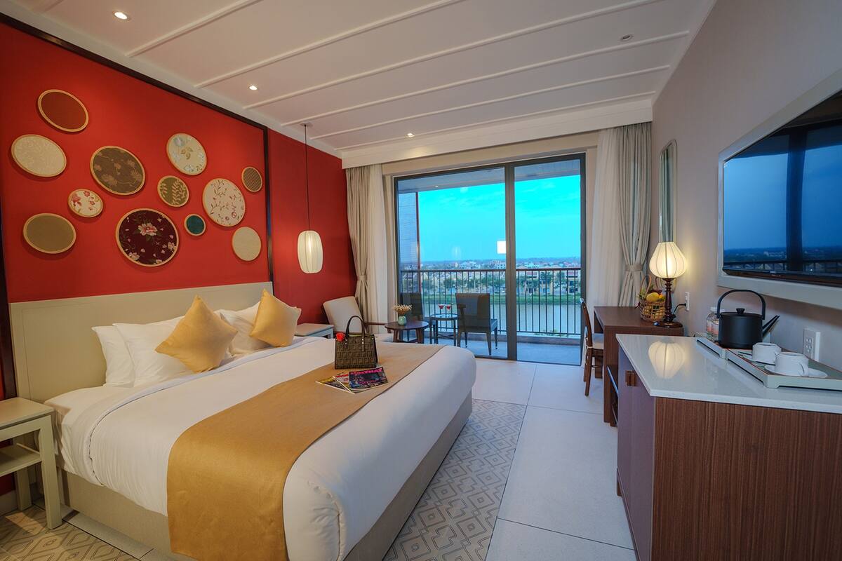 A hotel room with a large bed, a desk, and a balcony with a city view.