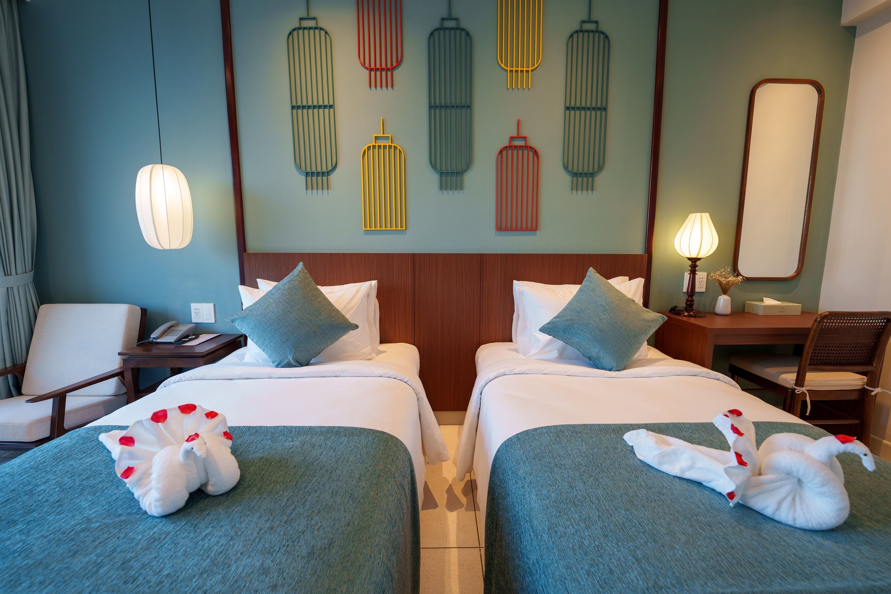 A hotel room with two beds, a desk, a chair, a lamp, and a wall with decorative birdcage art.