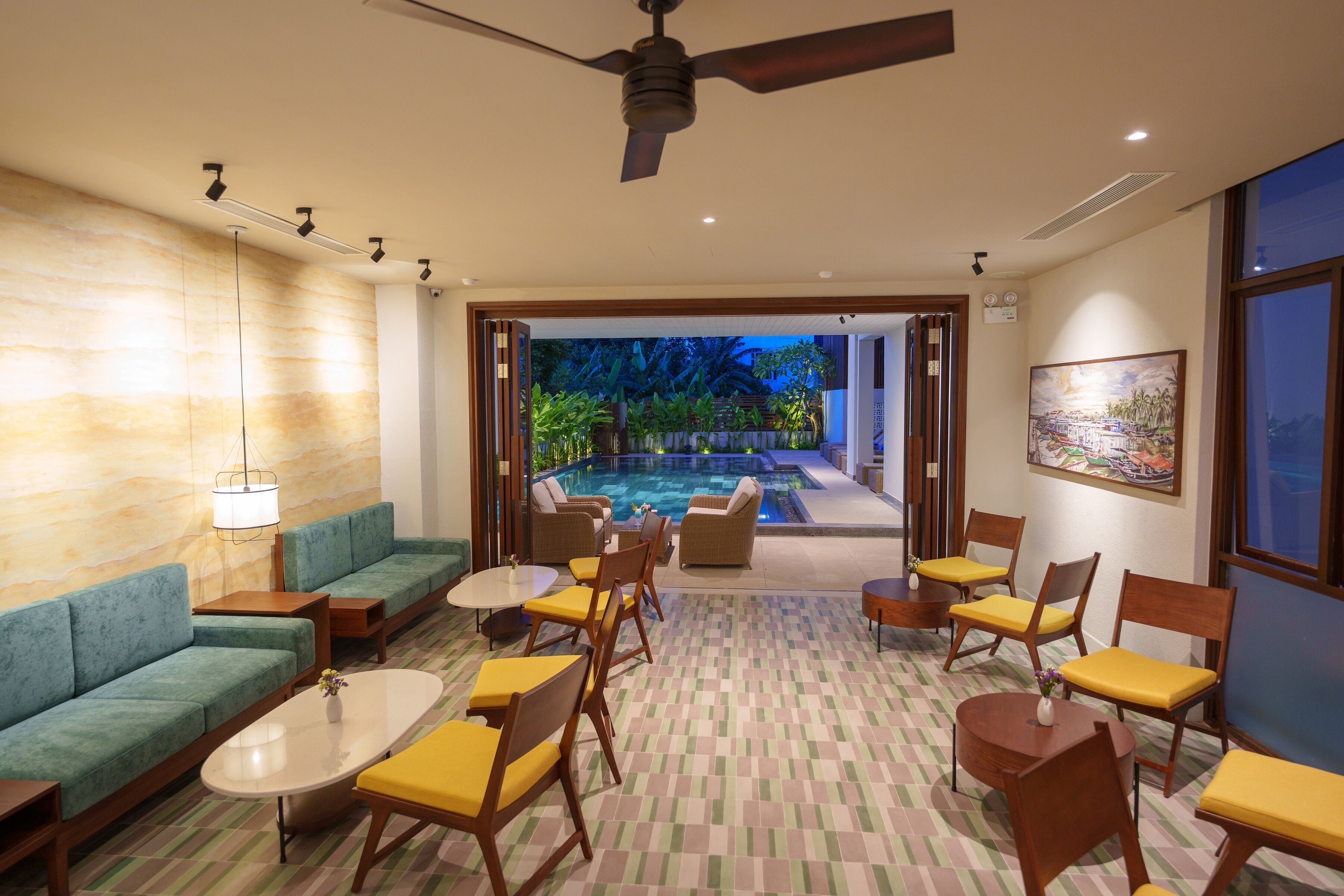 A hotel lobby with a pool view, green sofas, yellow chairs, and a ceiling fan.
