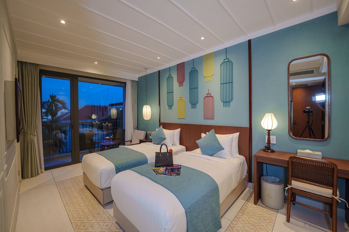 A hotel room with two beds, a desk, a chair, a waste bin, and a large window with a view of palm trees.