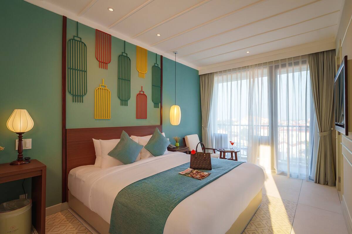 A hotel room with a large bed, a nightstand with a lamp, a chair, a TV, and a window with curtains.