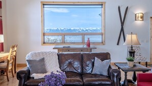 A cozy living room with a leather couch, a wooden dining table, and a large window overlooking a snowy mountain. - Spacious 5BR Mountain Retreat near Grand Targhee (Driggs)