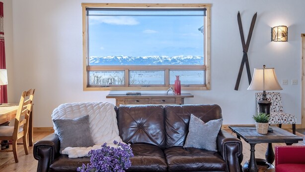 A cozy living room with a leather couch, a wooden dining table, and a large window overlooking a snowy mountain. - Spacious 5BR Mountain Retreat near Grand Targhee (Driggs)