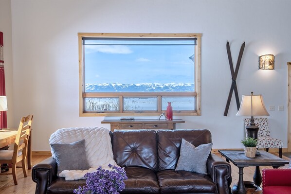 Een gezellige woonkamer met een leren bank, een houten eettafel en een groot raam met uitzicht op een besneeuwde berg.