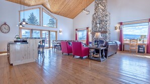 A spacious living area with wooden floors, a stone fireplace, large windows, and a dining area with red chairs. - Spacious 5BR Mountain Retreat near Grand Targhee (Driggs)