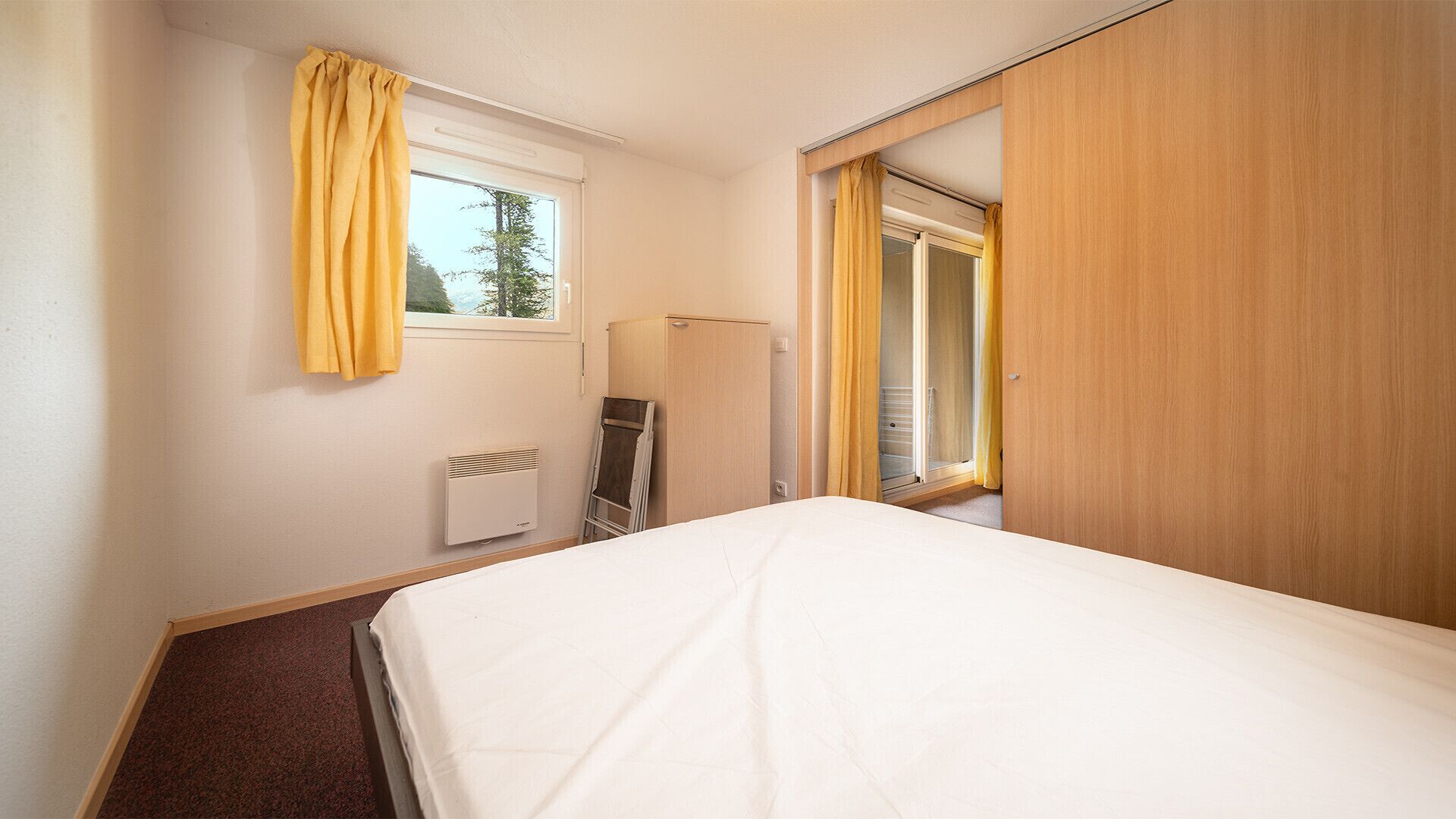 A bedroom with a bed, a radiator, a wardrobe, and a window with curtains.