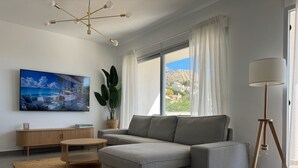 A modern living room with a grey sectional sofa, a wooden coffee table, a large flat-screen TV, a floor lamp, and a view of the outdoors through a large window. - ALTHAIA HILLS- Beautiful townhouse with panoramic terrace and sea and mountain views (Altea)