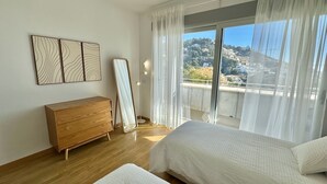 A bedroom with a wooden dresser, a standing mirror, two beds, and a view of a hillside through large windows. - ALTHAIA HILLS- Beautiful townhouse with panoramic terrace and sea and mountain views (Altea)