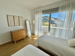 A bedroom with a wooden dresser, a standing mirror, two beds, and a view of a hillside through large windows.