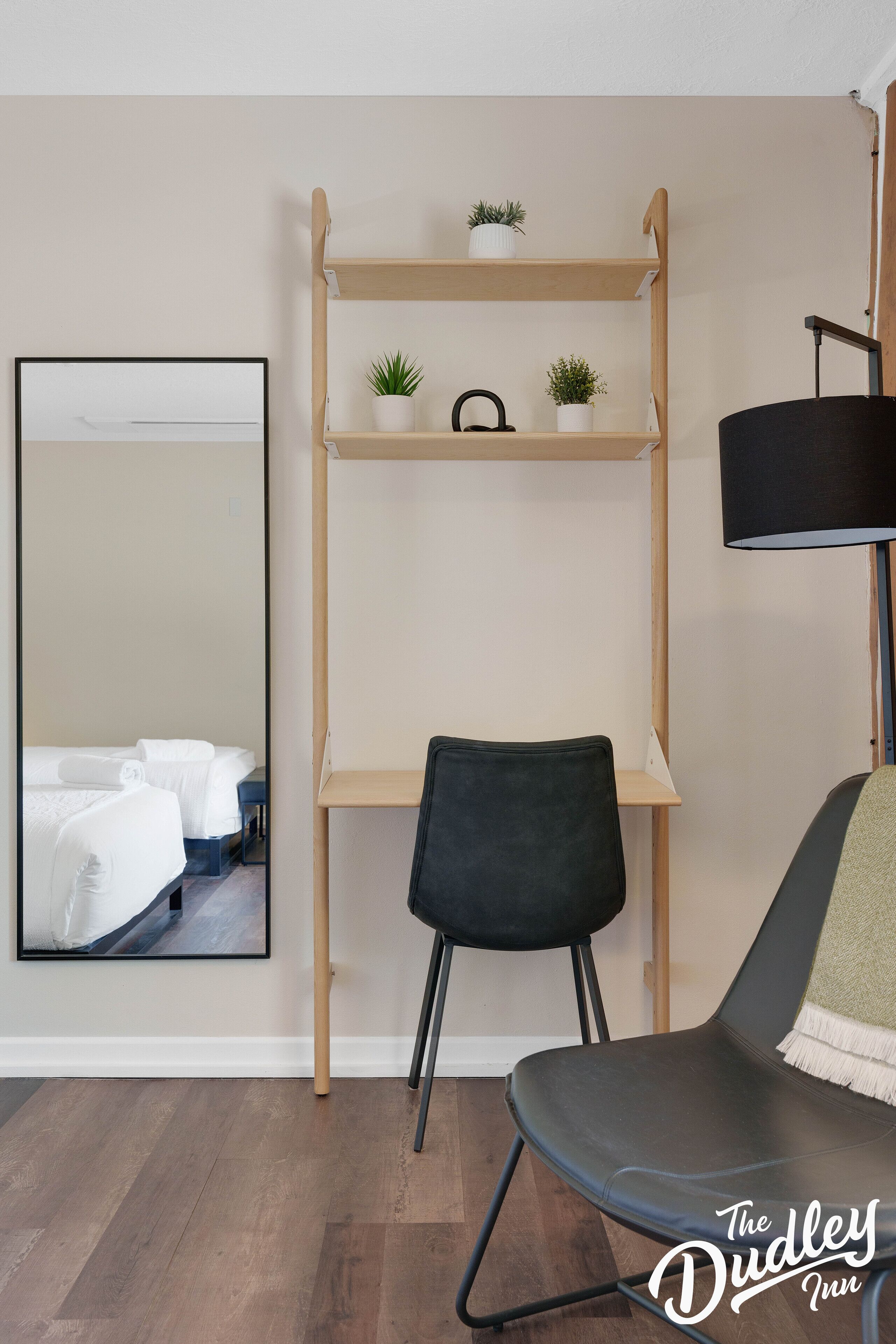 A room with a bed, a desk with a chair, a mirror, and a shelf with plants.