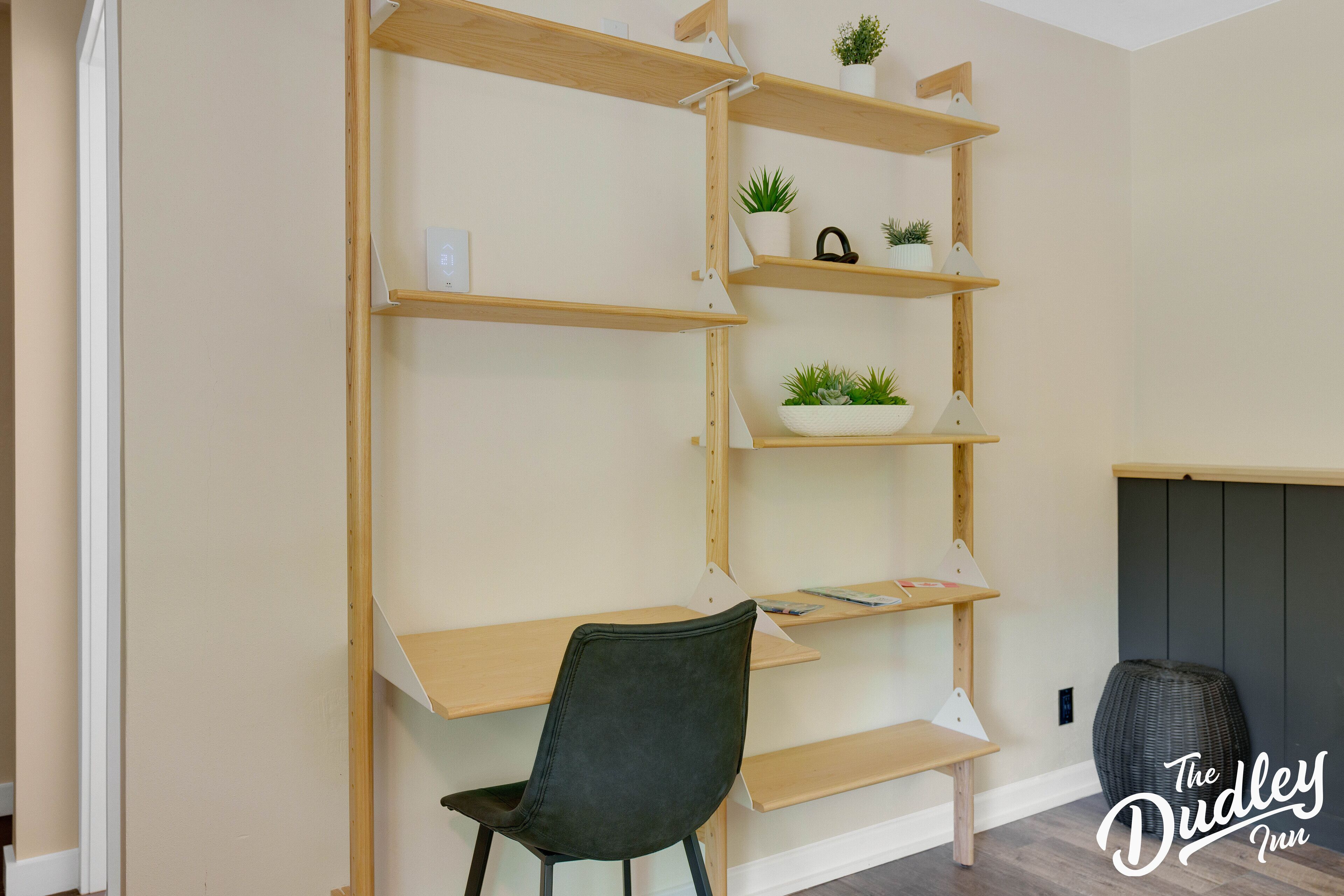 Una habitación con un escritorio de madera y estanterías, una silla negra y una planta en maceta sobre las estanterías.