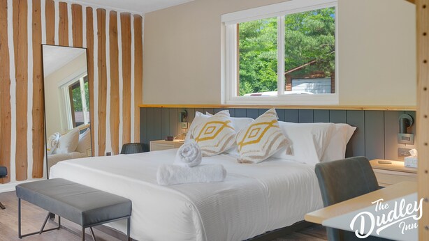 A hotel room with a large bed, a bench, a bedside table, a nightstand, a mirror, and a window with a view of greenery. - The Dudley Inn (Muskoka Lakes)