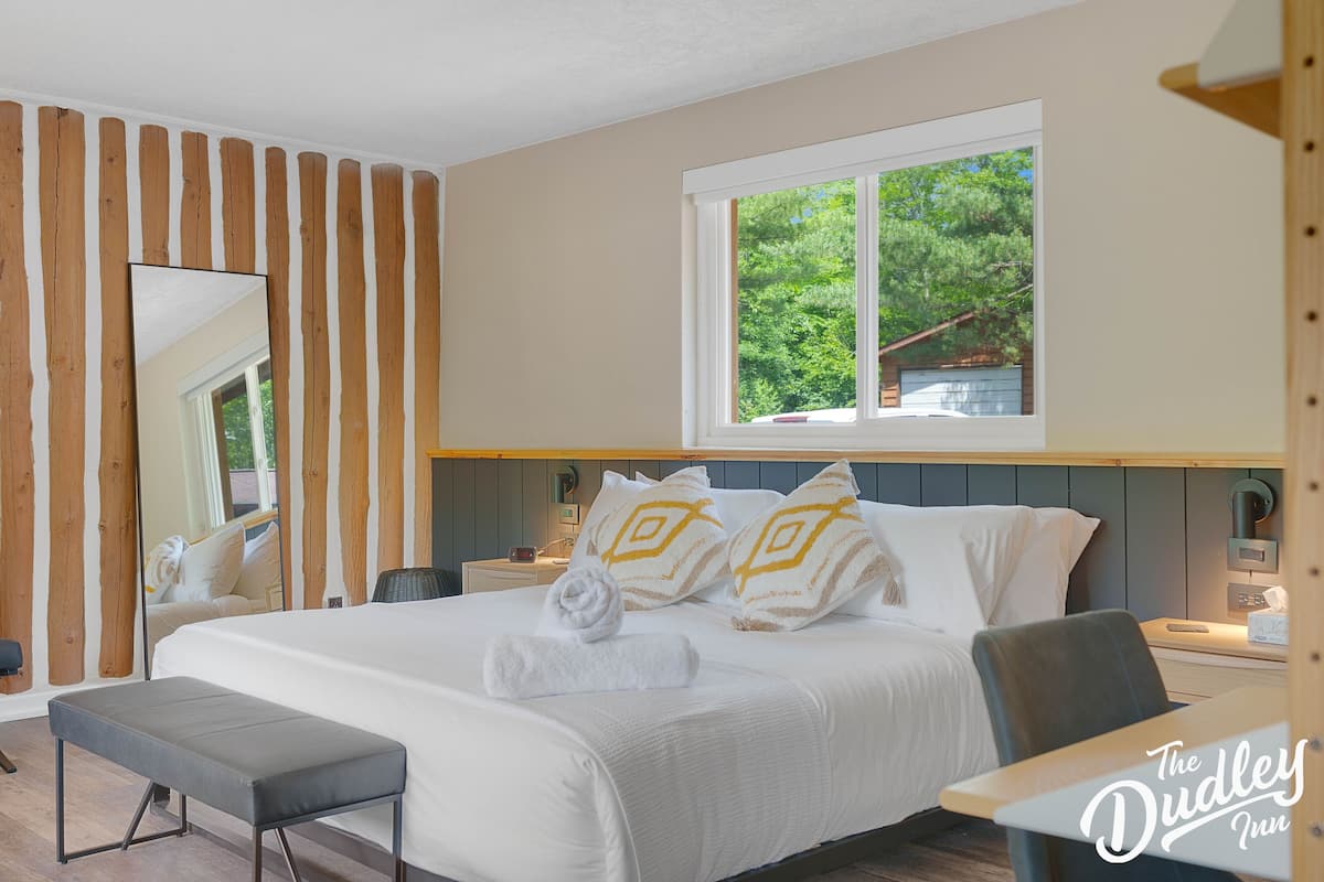 A hotel room with a large bed, a bench, a bedside table, a nightstand, a mirror, and a window with a view of greenery.