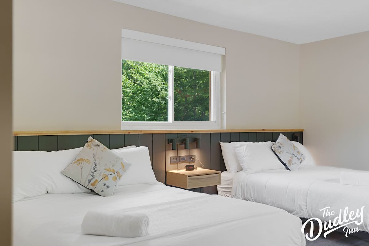 A hotel room with two beds, a nightstand with a clock, and a window with a view of greenery.