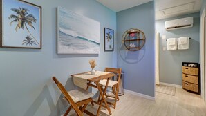 A dining area with a table and chairs, a wall-mounted shelf with decorative items, and a view of the beach through a window. - Private Surf Shack Less Than 5 Minutes to Indian Rocks Beach
Bikes Included! (Largo)